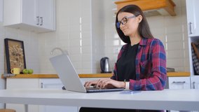 Young entrepreneur woman typing on laptop keyboard in 4k video. Pretty white female working on notebook pc in home kitchen during lockdown. Nerdy person wearing glasses and doing distant work online