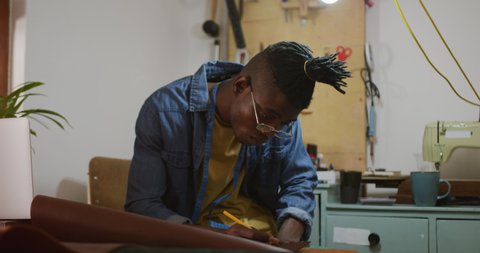 Thoughtful african american craftsman wearing glasses making notes in leather workshop. independent small business craftsman at work.