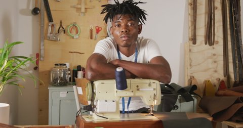 Thoughtful african american craftsman with dreadlocks leaning on sewing machine in leather workshop. independent small business craftsman at work.