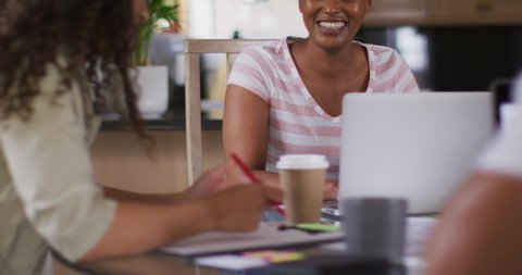 Two happy diverse female friends sitting in kitchen with coffee, working on laptop and laughing. technology and communication, flexible working from home.