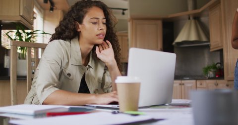 Thoughtful biracial woman sitting in kitchen and working on laptop. technology and communication, flexible working from home.