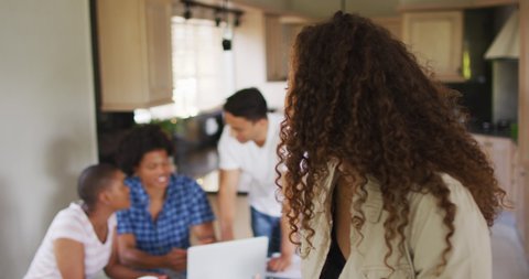 Happy biracial woman standing in kitchen and looking at camera with diverse friends in background. technology and communication, flexible working from home.