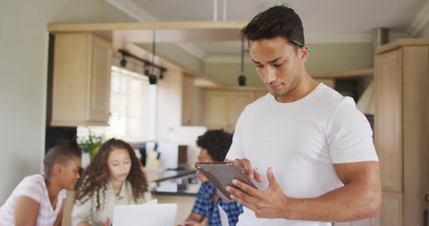 Happy latin man standing in kitchen with tablet and diverse friends in background. technology and communication, flexible working from home.