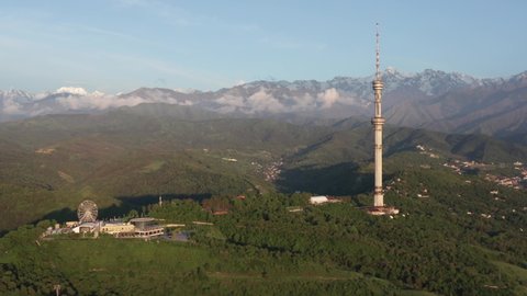 Almaty, Kazakhstan - June 29, 2020:
Aerial footage of Almaty city TV tower at summer sunset. Green Almaty city hills with clouds above. TV tower in the mountains wide shot.