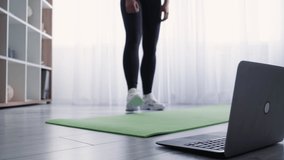 Online fitness training. Sportive woman. Warming exercise. Healthy lifestyle. Unrecognizable athletic lady in sportswear and shoes doing stretching workout footstep before laptop light room interior.