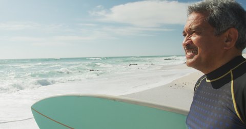 Portrait of happy senior hispanic man walking on beach with surfboard. sporty, healthy and active retirement lifestyle.