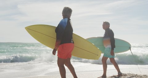Side view of happy senior hispanic couple walking on beach with surfboards. sporty, healthy and active retirement lifestyle.