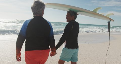 Back view of happy senior hispanic couple walking on beach with surfboard. sporty, healthy and active retirement lifestyle.