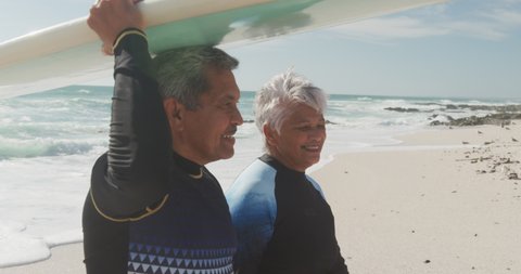 Profile of happy senior hispanic couple walking on beach with surfboard. sporty, healthy and active retirement lifestyle.