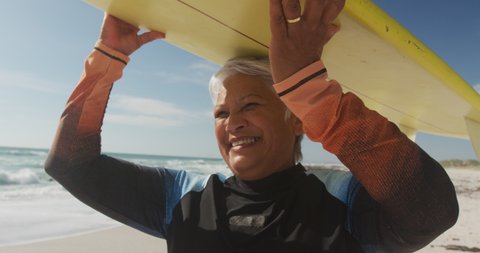 Profile of happy senior hispanic woman walking on beach with surfboard over head. sporty, healthy and active retirement lifestyle.