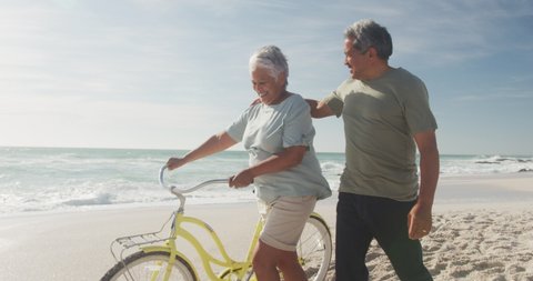 Happy hispanic senior couple walking with bikes on beach at sunset. sporty, healthy and active retirement lifestyle.