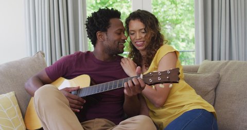 Happy biracial couple sitting on sofa together and playing guitar. love, relationship and spending quality leisure time at home.