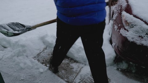 A man in a blue jacket and black pants is shoveling snow outside his house. A red car is standing nearby. Snow is falling from the sky