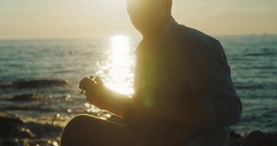 Fun attractive man traveler sitting on sea beach with ukulele under sunset sky in evening time. Male playing, sing music, relaxing enjoying holidays, travel day. 4K video