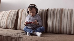 A 4-year-old boy in blue jeans and a gray turtleneck in headphones sits on the couch and looks enthusiastically at the phone