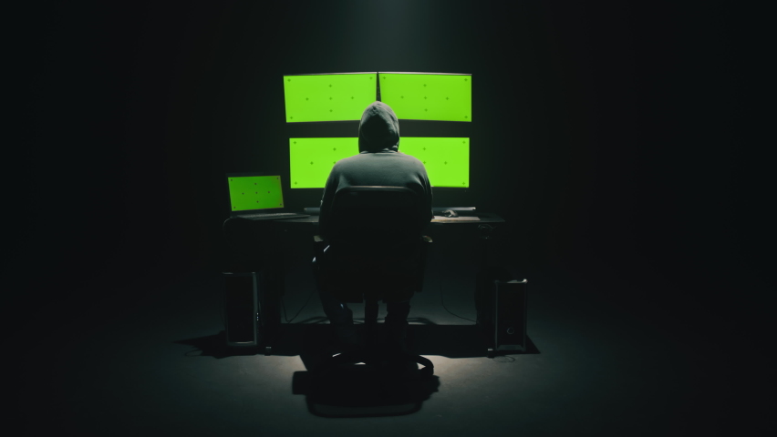 Rear view of hooded anonymous scammer hacker at table with green screen chromakey multiple display, during an attack on credit cards big data servers on the network Royalty-Free Stock Footage #1086911609