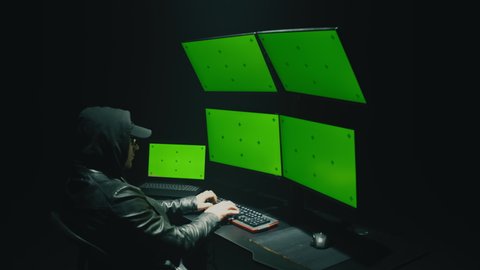 A male cybercriminal in glasses and a hood hacking the security system on the network and infecting with a virus in data on a computer with software, in a dark room at night