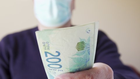 Giving cash Sheqel Israel currency paper banknotes. Closeup of israeli man in Protective face mask paying bills during pandemic