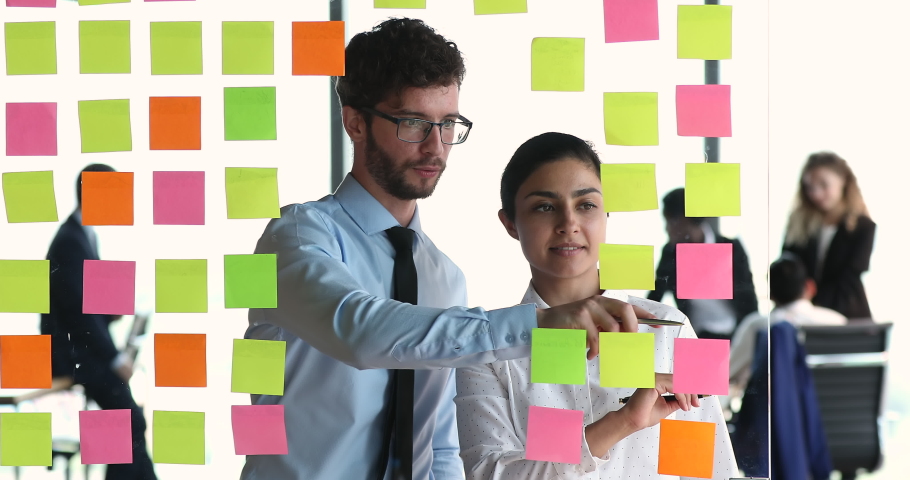 Young coworkers program code writers create software interface structure place colored post it paper sheets on glass wall. Two mates managers Indian woman European man talk make marks on kanban cards Royalty-Free Stock Footage #1086926438