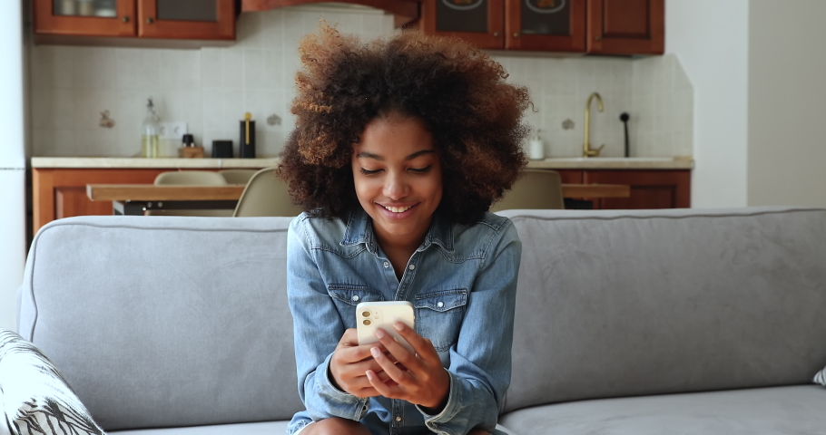 Beautiful African teenage girl sit on sofa holding cell phone texting sms, share messages in social media, communicates remotely use wireless gadget. Young gen Z and modern technology usage concept Royalty-Free Stock Footage #1086926465