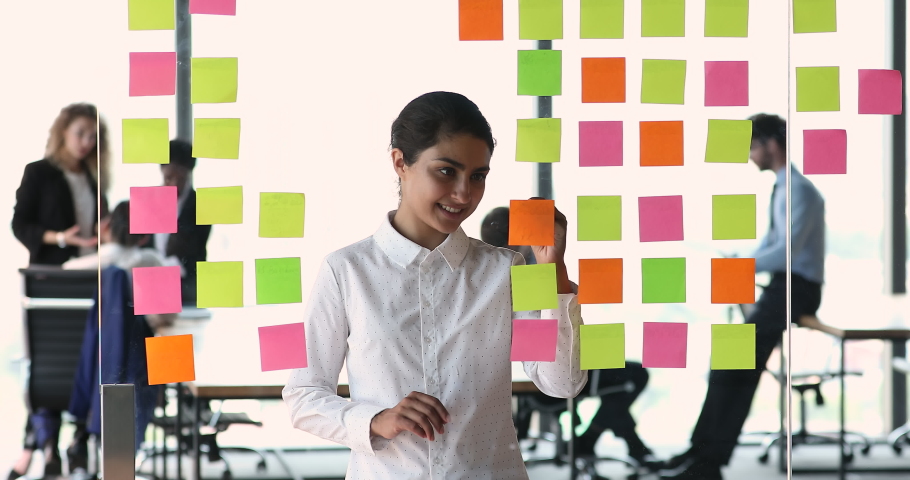 Smiling Indian businesswoman project manager write compare data on colored stickers based on glass board. Young lady software engineer work with information develop new task using kanban or scrum tool Royalty-Free Stock Footage #1086926495