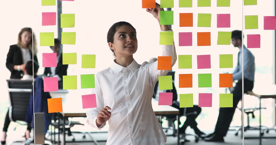 Smiling Indian businesswoman project manager write compare data on colored stickers based on glass board. Young lady software engineer work with information develop new task using kanban or scrum tool | Shutterstock HD Video #1086926495