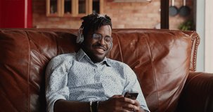 Smiling African man in wireless earphones relax on sofa using phone audio app watch movie video clip select music track in online playlist. Happy male hipster spend free time playing web game on cell