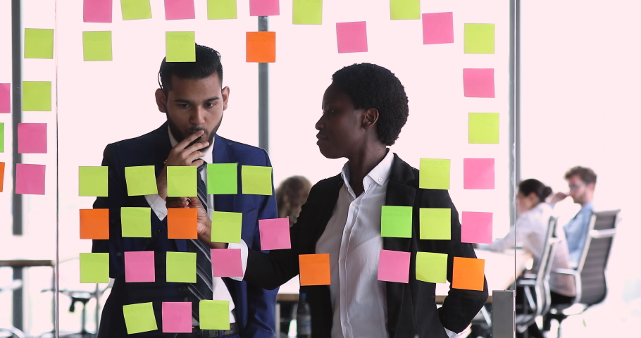 Diverse business partners share ideas stand near glass wall with structured business process model. Black woman Caucasian man teammates discuss project vision analyze benefits using paper memo sticks Royalty-Free Stock Footage #1086926672