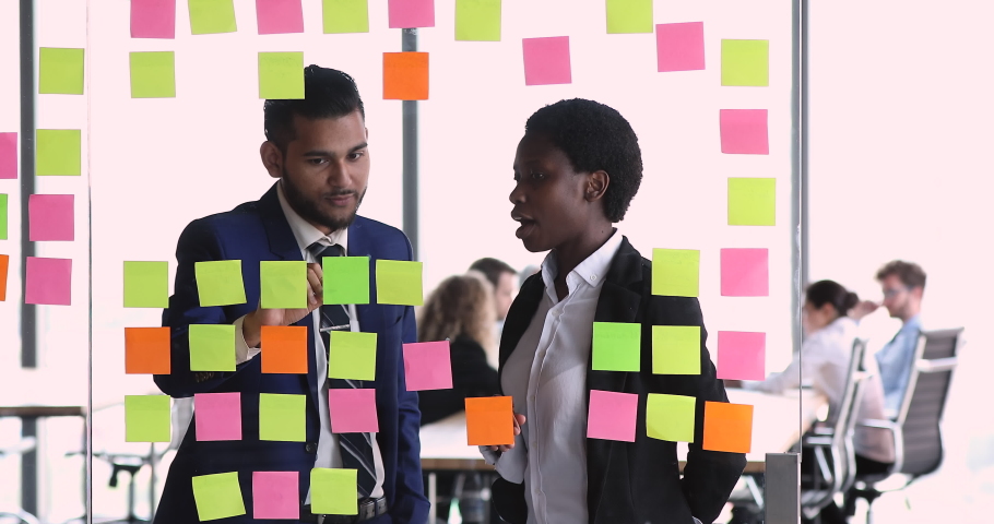 Diverse business partners share ideas stand near glass wall with structured business process model. Black woman Caucasian man teammates discuss project vision analyze benefits using paper memo sticks | Shutterstock HD Video #1086926672