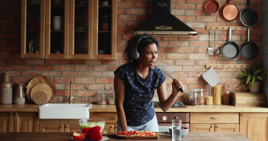 Energetic African female wear wireless headset imitate karaoke sing song preparing lunch pretend singer using whisk as microphone. Funny young woman enjoy easy cooking food at modern designed kitchen Royalty-Free Stock Footage #1086926681