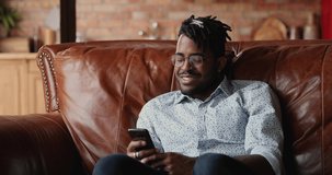 Glad Black male hipster rest on couch use phone at leisure time buy goods online read funny message in web chat . Positive young man with stylish dreadlocks watch video content at social media on cell