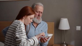 Side view of happy redhead granddaughter showing photo using mobile photo to gray-haired grandfather sitting on bed at home. Attractive young woman teaching mature adult male how to use smartphone.
