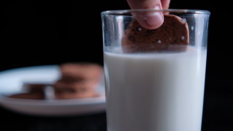 Hand slowly grabbing chocolate chip cookie and dunking it in glass of milk. Close-up of fresh milk and white plate of tasty cookies with pitch-black background. Sweet food and snacks