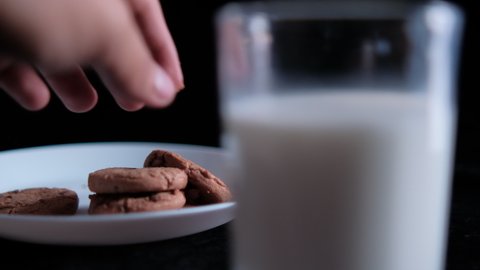Hand slowly grabbing chocolate chip cookie from plate behind blurry glass of milk. Close-up of fresh milk and white plate of tasty cookies with pitch-black background. Sweet food and snacks