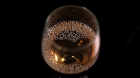 Top view of bubbly beer in glass goblet with pitch-black background. Smooth view of golden frothy alcoholic drink in gleaming wine glass on black surface. Beverages and celebrations