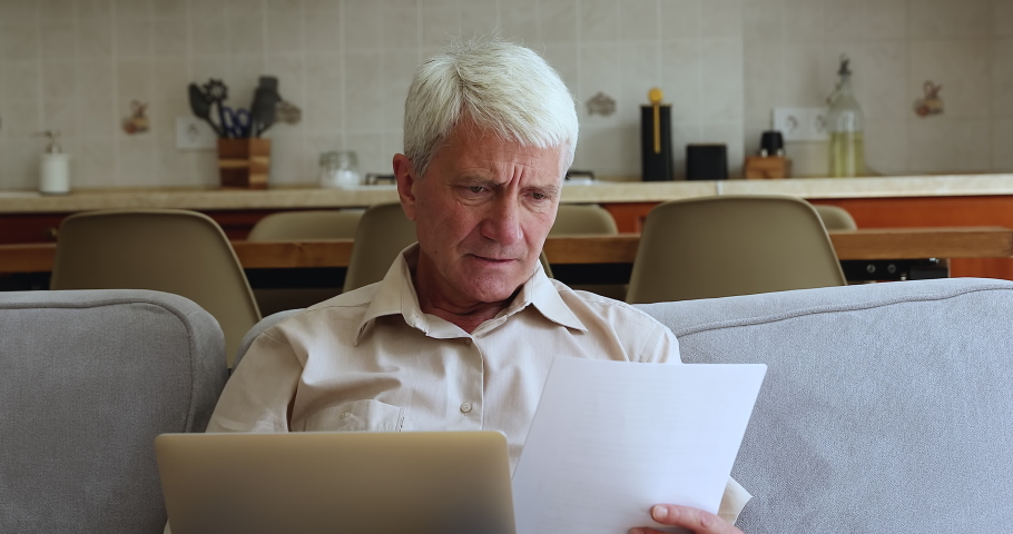Angry older man sits on sofa with laptop hold paper, read notice feels negative emotions looks annoyed by mistake in bank document, overspend, lack of finance to pay huge taxes or bills, debt concept Royalty-Free Stock Footage #1086932924