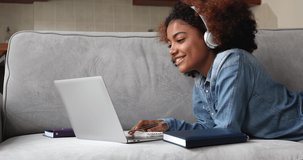 African teenage girl lying on couch studying from home use computer and video call application, take notes, do college assignment, prepare for university admission or exams. E-learning, tech concept