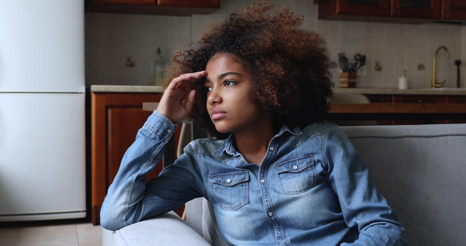 Sad African teenage girl sit on sofa at home, lost in thoughts, looks into distance, think, looks upset experiences first unrequited love, having problems in school feel insecure. Teen problem concept | Shutterstock HD Video #1086932963