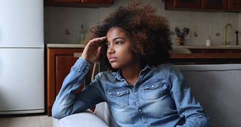 Sad African teenage girl sit on sofa at home, lost in thoughts, looks into distance, think, looks upset experiences first unrequited love, having problems in school feel insecure. Teen problem concept