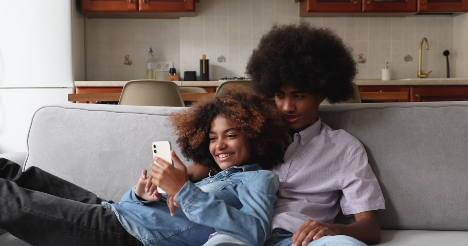 Beautiful African teenage couple relaxing on sofa with gadget, use new mobile application, enjoy cool filter have fun use smartphone spend time together indoor. Young gen and modern technology concept Royalty-Free Stock Footage #1086933002