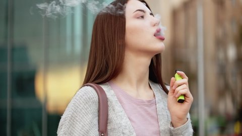 Vaping woman. Electronic cigarette. Nicotine addiction. Attractive elegant young female smoking outdoors exhaling steam at city street.