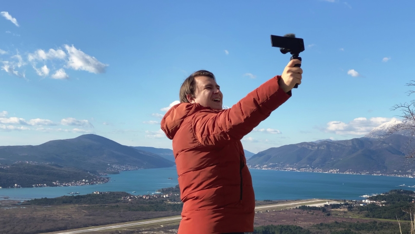 Man tourist in jacket records himself talking on camera against sea Bay of Kotor and forestry mountains IN old town of Montenegro