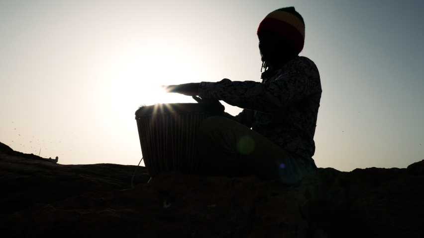 African percussion music, silhouette of a Senegalese man playing a traditional djembe drum with his hands at sunset. Outdoor music at the beach of Essaouira, Morocco. 4k
