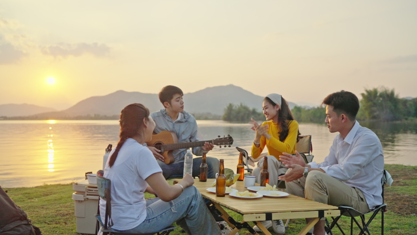 Group Four People Friends Asian men and women Camping, drinking beer, celebrating, Singing, Playing Guitar, Having fun and Enjoying Ground tent. Reservoir area during Sunset Vacation Time.