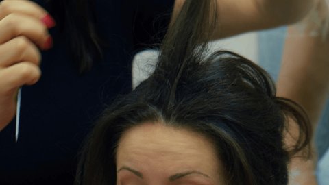 Hairstyling with hair spray, preparation for prom. Close-up view of stylist hairdresser making hairstyle using curling tongs for long hair of unrecognizable woman in beauty salon.