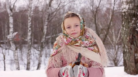 Cute girl in a traditional Russian headscarf and mittens playing on spoons for Maslenitsa festival on winter background. Closeup portrait of a child for Srovetide holiday.