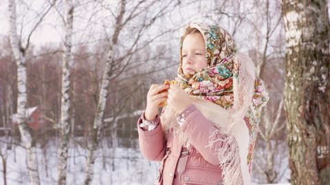 Cute girl in a traditional Russian headscarf with bagels eats pancake on winter background. Closeup portrait of a child in folk clothes. Maslenitsa festival. Traditional Russian food for Shrovetide.