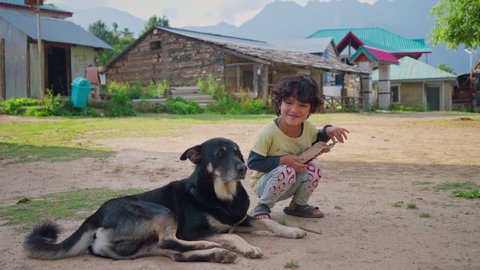 A shot of a street dog and a young cute Asian Indian little rural girl sitting together on the ground looking at camera with Traditional Village houses in the background in a mountain region