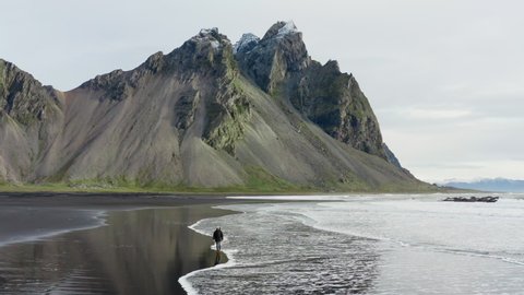 Drone Shot Of Man On Black Sand Beach Walking As The Tide Comes In And Stopping To Look At View With Vestrahorn Mountain In The Background, Iceland