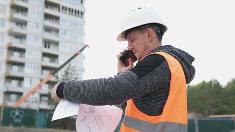 The foreman at the construction site of a high-rise building is talking on the phone and checking the drawings.